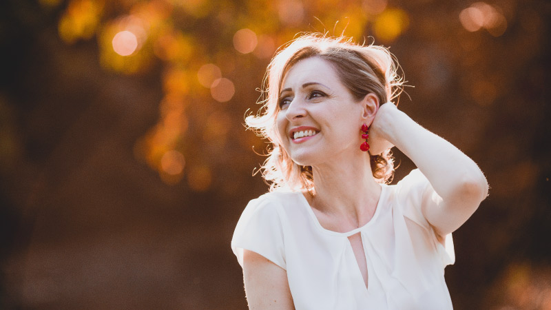 Smiling blonde woman wears a white blouse outdoors in front of golden brown leaves