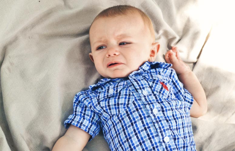 Cranky baby wearing a checkered blue button up shirt lies on a blanket while fussing due to teething pain