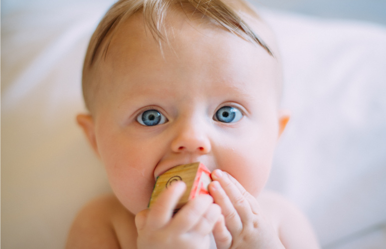 Blonde baby with bright blue eyes sticks a block into his mouth to ease his inflamed gums due to teething pain