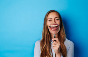 Brunette woman magnifies her teeth with a magnifying glass against a blue wall