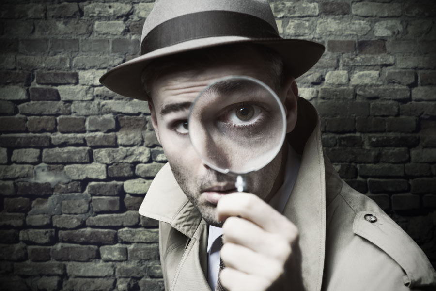 man in a hat and trench coat holds a magnifying glass up to his eye 