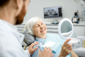 senior woman holds a mirror to look at her teeth while sitting in the dentist chair with the dentist standing by